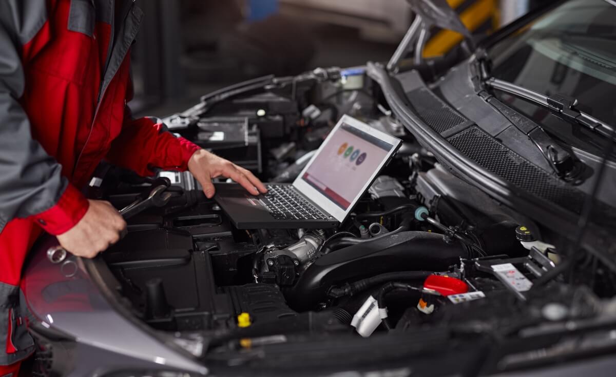 Mechanik pracuje przy otwartej masce samochodu, używając laptopa do diagnostyki komputerowej silników.